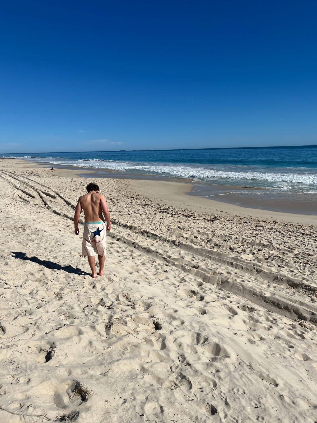 A man walks away on a sandy beach wearing light-colored swim trunks with a dark graphic print, with the ocean and blue sky in the background.
