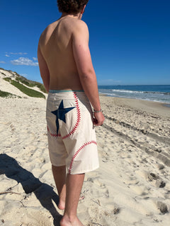 A shirtless man standing on a sandy beach, viewed from behind, wearing cream baseball-themed board shorts with red stitching and a navy blue star graphic.