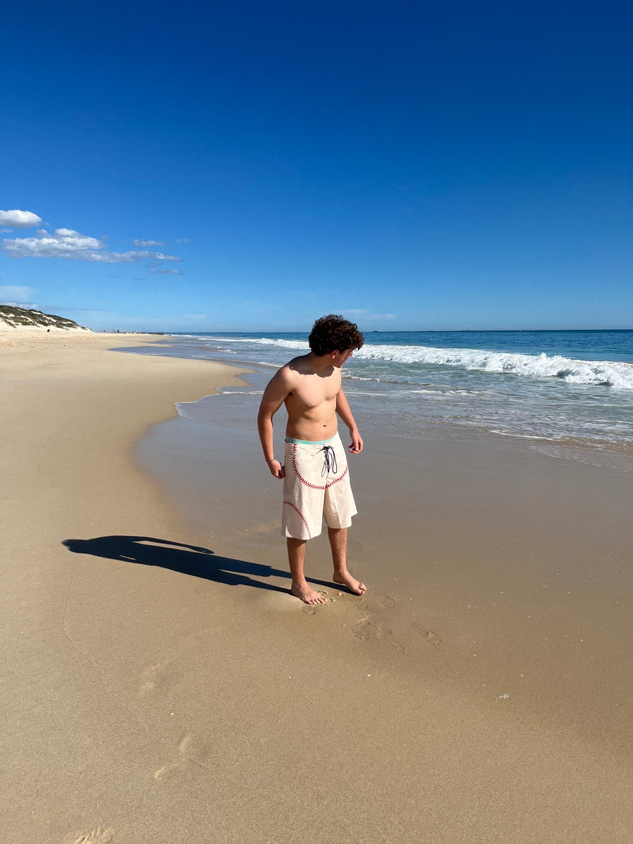 A shirtless man standing on a sandy beach near the shoreline, wearing light cream swim trunks with a navy drawstring. The background features a clear blue sky, ocean waves, and a long shadow cast on the sand.